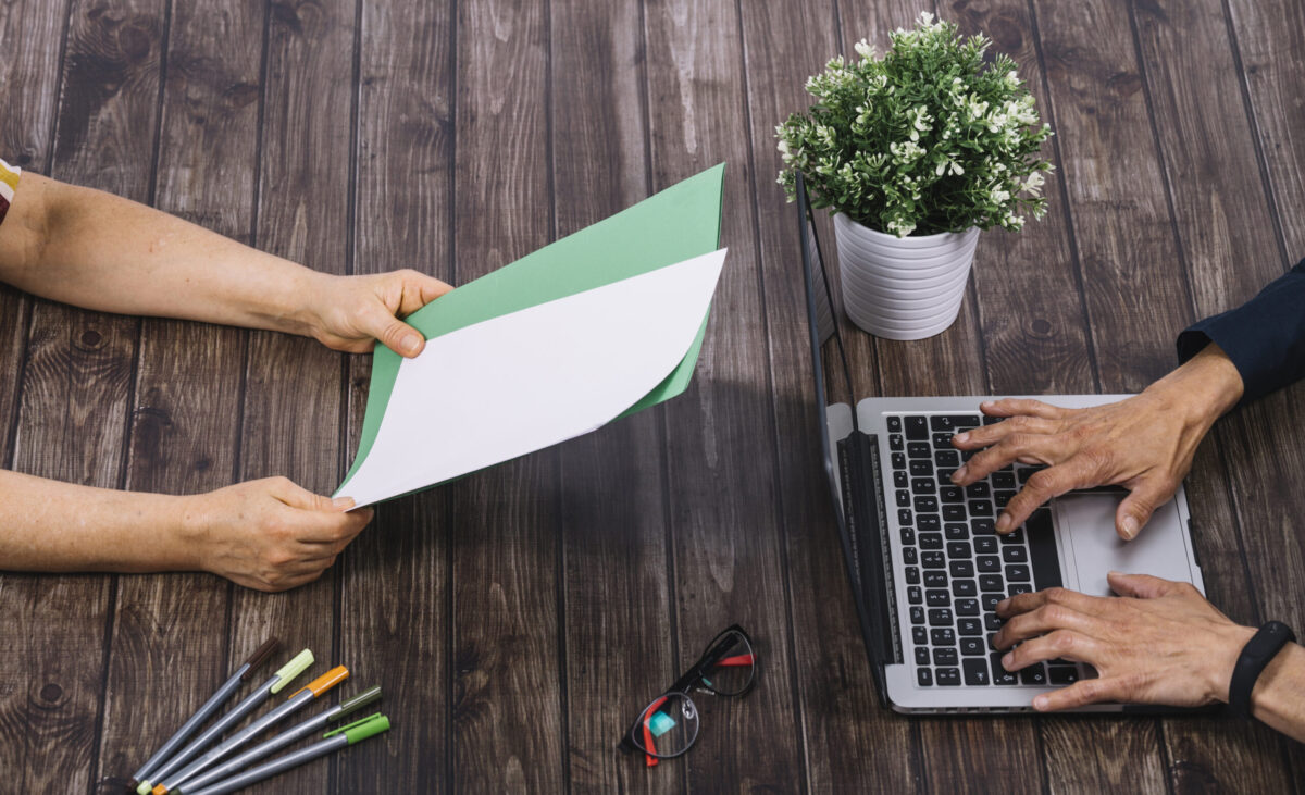 person-typing-laptop-with-his-colleague-holding-blank-white-green-blank-paper-wooden-table person-typing-laptop-with-his-colleague-holding-blank-white-green-blank-paper-wooden-table